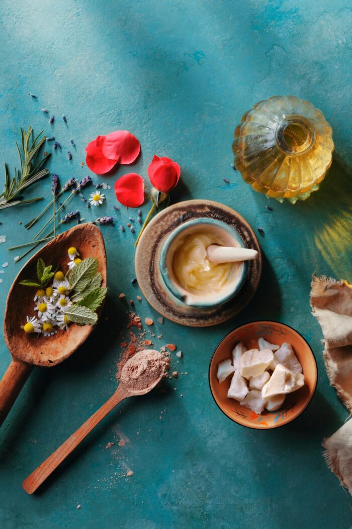 Overhead flat lay of botanical skincare ingredients including rose petals, chamomile, lavender, and oils on a teal textured surface.